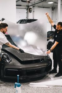 Two technicians applying a protective film on a sleek sports car in a garage setting.
